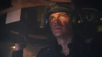 Auf dem Bild ist David Schütter in seiner Rolle im Film „Der Tiger“ zu sehen. Er befindet sich im Inneren eines Panzers, trägt Kopfhörer und eine Uniform mit Mütze. Sein Gesicht wird von einem warmen Licht angestrahlt, wodurch seine angespannte und konzentrierte Mimik noch deutlicher wirkt. Der enge Raum im Panzer und die Schatten um ihn herum unterstreichen die beklemmende Atmosphäre der Szene. - Foto: Amazon MGM Studios