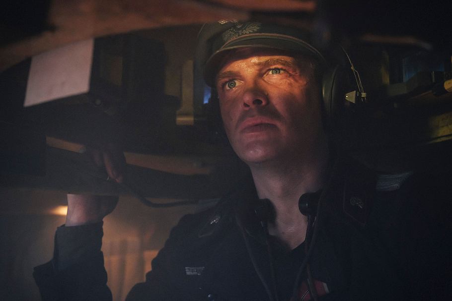 Auf dem Bild ist David Schütter in seiner Rolle im Film „Der Tiger“ zu sehen. Er befindet sich im Inneren eines Panzers, trägt Kopfhörer und eine Uniform mit Mütze. Sein Gesicht wird von einem warmen Licht angestrahlt, wodurch seine angespannte und konzentrierte Mimik noch deutlicher wirkt. Der enge Raum im Panzer und die Schatten um ihn herum unterstreichen die beklemmende Atmosphäre der Szene.