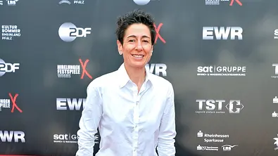 Dunja Hayali steht lächelnd in weißem Hemd und blauer Hose auf einem roten Teppich vor einer Wand mit Sponsorenlogos. - Foto: IMAGO / STAR-MEDIA
