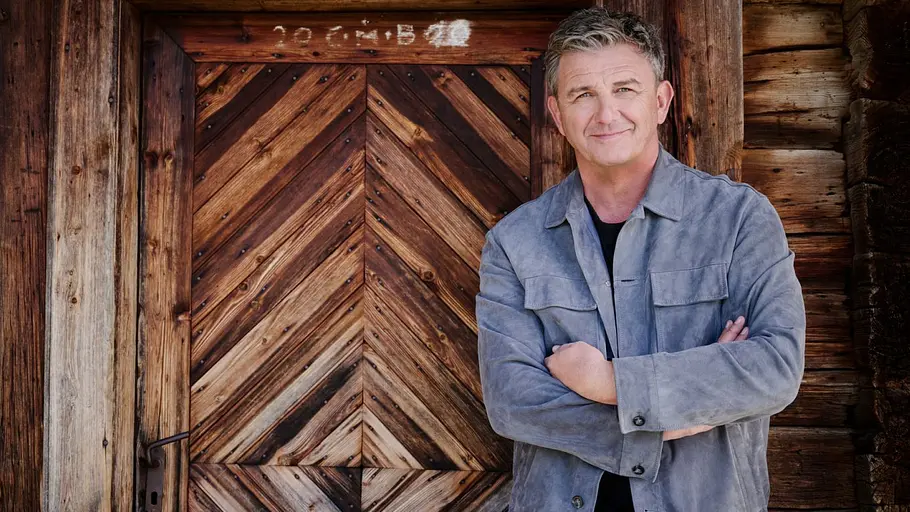 Hans Sigl steht vor einer Holzhütte und verschränkt lächelnd die Arme. 