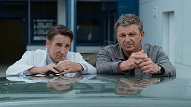 Die Arme auf das Autodach abgelegt stehen Dr. Alexander Kahnweiler (Mark Keller) und Dr. Martin Gruber (Hans Sigl) nebeneinander. - Foto: ZDF und Sabine Finger Fotografie. Die Arme auf das Autodach abgelegt stehen Dr. Alexander Kahnweiler (Mark Keller) und Dr. Martin Gruber (Hans Sigl) nebeneinander. - Foto: ZDF und Sabine Finger Fotografie.