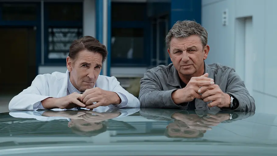 Bergdoktor Die Arme auf das Autodach abgelegt stehen Dr. Alexander Kahnweiler (Mark Keller) und Dr. Martin Gruber (Hans Sigl) nebeneinander.