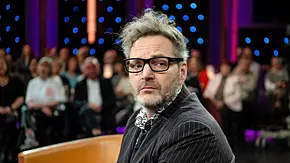 Paul Panzer sitzt mit schwarzer Brille, Headset und Nadelstreifenanzug vor einem Publikum im bunt beleuchteten Studio. - Foto: Imago / STAR-MEDIA Paul Panzer sitzt mit schwarzer Brille, Headset und Nadelstreifenanzug vor einem Publikum im bunt beleuchteten Studio. - Foto: Imago / STAR-MEDIA