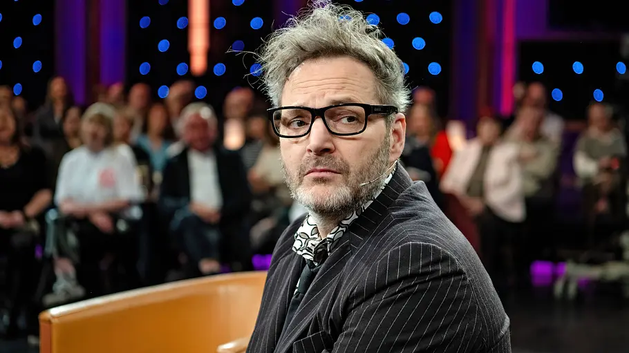 Der Grund für Paul Panzers Rückzug war lange unbekannt. Paul Panzer sitzt mit schwarzer Brille, Headset und Nadelstreifenanzug vor einem Publikum im bunt beleuchteten Studio.