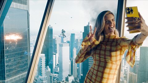 Eve Teschmacher macht ein Selfie vor einem Fenster, hinter ihr findet ein Kampf im Himmel statt. - Foto: Warner Bros.