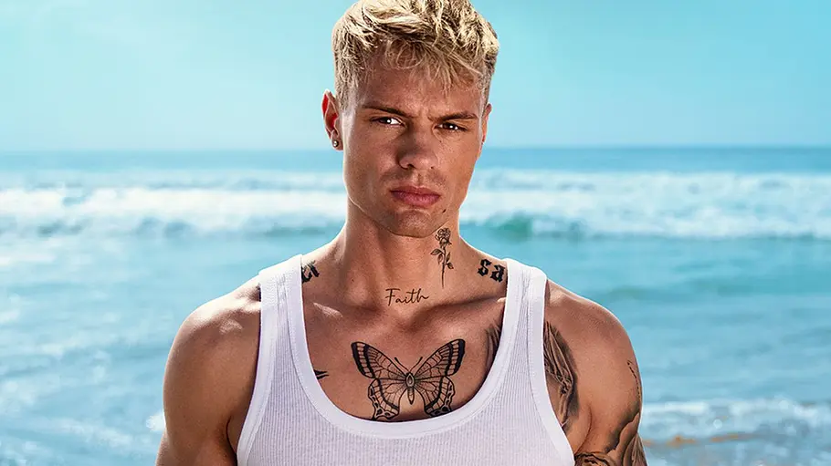 „Der Promihof“-Kandidat Fabian Hesdahl Fabian Hesdahl steht mit blondem Haar, weißem Tanktop und Tattoos an Hals, Brust und Armen vor dem Meer.