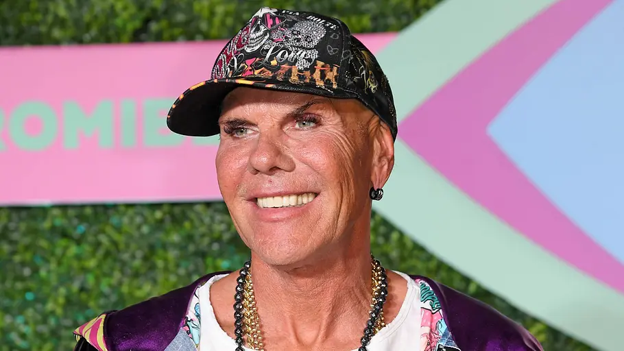 „Der Promihof“-Kandidat Redo Ein lächelnder Mann trägt eine bunt bedruckte Kappe, Ketten und eine glänzende Jacke vor einem grün-rosa Hintergrund.