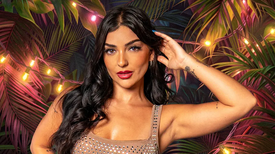 „Der Promihof“-Kandidatin Jennifer Iglesias Jennifer Iglesias posiert im beigen, glitzernden Kleid mit einer Hand am Kopf vor tropischen Blättern und bunten Lichterketten.