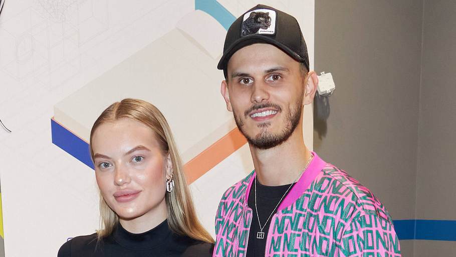 Cheyenne Ochsenknecht steht neben ihrem Mann und Moderator von der Promihof Nino Sifkovits. Beide lächeln in die Kamera.