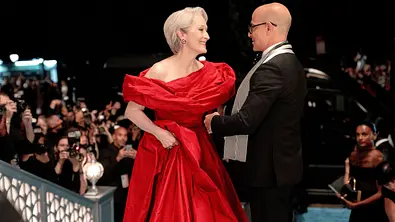 Miranda im roten Abendkleid wird begrüßt von Nigel. Rundherum steht die Presse, alle Kameras sind auf sie gerichtet. - Foto: 2026 20th Century Studios