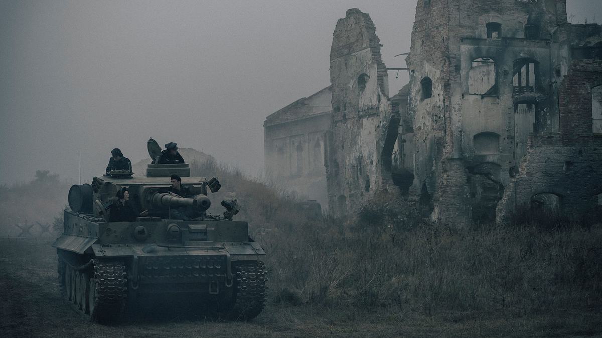 Ein schwerer Panzer vom Typ Tiger rollt auf einem matschigen Weg durch eine graue, neblige Landschaft. Im Hintergrund ragen die Ruinen eines zerbombten Gebäudes auf, ausgebrannte Fensterhöhlen und zerfallene Mauern. Zwei Soldaten sind oben aus den Luken des Panzers zu sehen.