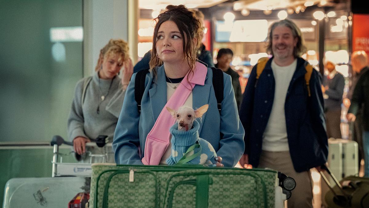 Der Umzug nach London hat sich für Jess gelohnt. Jess mit Koffern und Hund am Flughafen. Sie schaut skeptisch zur Seite.