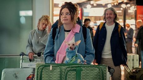 Jess mit Koffern und Hund am Flughafen. Sie schaut skeptisch zur Seite. - Foto: © 2024 Netflix, Inc.