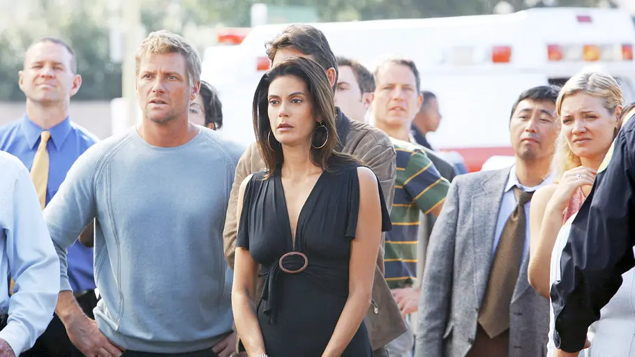 Desperate Housewives Susan und Tom beobachten gemeinsam mit mehreren Nachbarn und Einsatzkräften ein dramatisches Geschehen auf der Straße. Im Hintergrund ist ein Rettungswagen zu sehen – Szene aus „Desperate Housewives“.