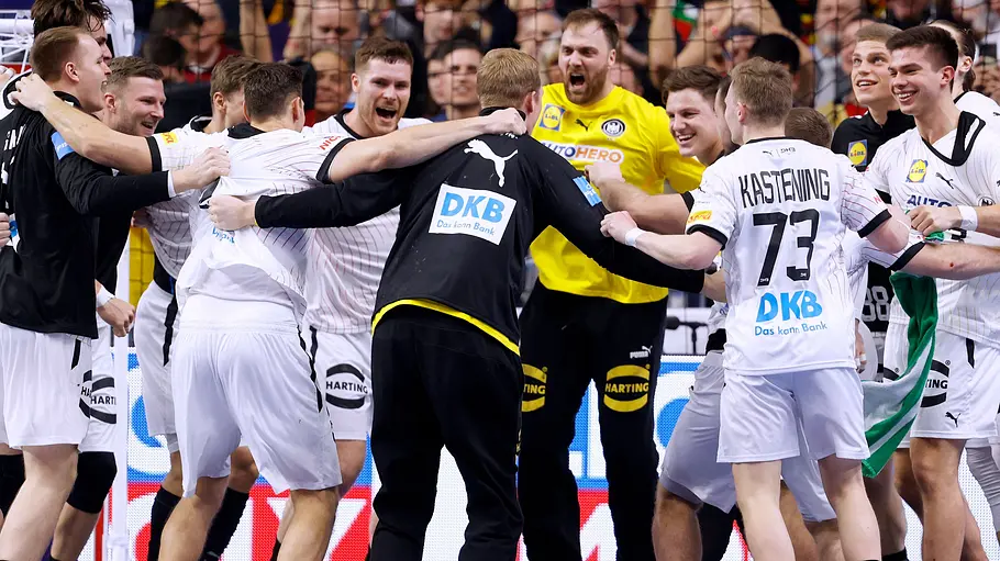 Die deutsche Handball-Nationalmannschaft jubelt gemeinsam im Kreis vor dem Tor. Auf der Tribüne stehen zahlreiche Zuschauer.
