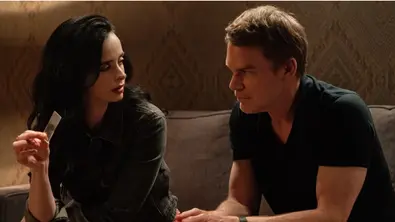 Dexter Morgan sitzt neben einer dunkelhaarigen Frau auf einem Sofa. Sie hält ein kleines rechteckiges Plättchen in der Hand. - Foto: Matt Infante/Paramount+ Dexter Morgan sitzt neben einer dunkelhaarigen Frau auf einem Sofa. Sie hält ein kleines rechteckiges Plättchen in der Hand. - Foto: Matt Infante/Paramount+