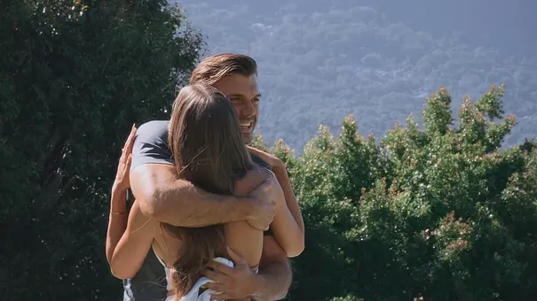Bachelor Tim und eine Kandidatin umarmen sich herzlich im Freien. Im Hintergrund sind grüne Bäume und bewaldete Berge zu sehen. Die Frau ist dem Betrachter abgewandt, der Mann lächelt glücklich. - Foto: RTL