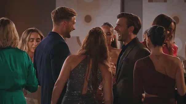 Die Bachelors und die Kandidatinnen stehen bei einer Abendveranstaltung in einem warm beleuchteten Raum zusammen. Sie lachen und unterhalten sich. - Foto: RTL
