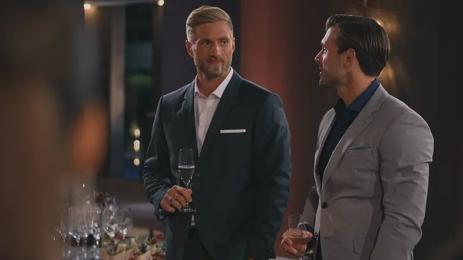 Die Bachelors Sebastian und Tim Sebastian und Tim im Anzug stehen bei einem Empfang nebeneinander, halten jeweils ein Glas in der Hand und unterhalten sich.
