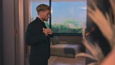 Bachelor Sebastian in einem schwarzen Anzug steht in einem modern eingerichteten Raum vor einem großen Fenster und richtet mit beiden Händen sein Jackett. - Foto: RTL Bachelor Sebastian in einem schwarzen Anzug steht in einem modern eingerichteten Raum vor einem großen Fenster und richtet mit beiden Händen sein Jackett. - Foto: RTL