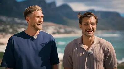 Zwei lächelnde Männer stehen nebeneinander vor einer Küstenlandschaft mit Bergen und Meer im Hintergrund und tragen Ansteckmikrofone, offenbar bei Dreharbeiten im Freien. - Foto: RTL