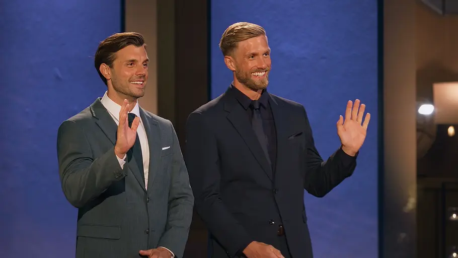 Die Bachelors Die Bachelors Tim und Sebastian stehen in Anzügen nebeneinander und Winken lächelnd in eine Richtung.