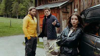 Die Bergretter: Vertrauen: Auf Emilies Hof: Eine zornig wirkende Mia Steiner (Lisa Junick) lehnt, die Arme vor der Brust verschränkt, an einem Auto und blickt in die Kamera. Im Hintergrund stehen Markus Kofler (Sebastian Ströbel) und Lorenz König (Steffen Groth) und unterhalten sich. Lorenz zeigt dabei mit der Hand auf Mia. - Foto: ZDF/Sabine Finger Fotografie