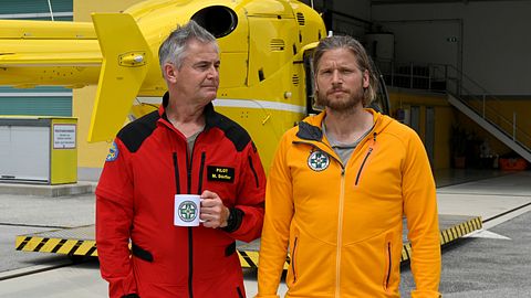 Zu sehen sind zwei Männer, die in einer roten Jacke (links) und einer orangenen Jacke vor einem gelben Hubschrauber stehen. Der Mann in der roten Jacke hält eine weiße Kaffeetasse in der Hand. - Foto: Media Press