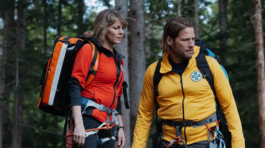 Katharina und Markus Katharina und Markus stehen in orange-roter und gelber Jacke mit Rucksäcken und Kletterausrüstung im Wald.