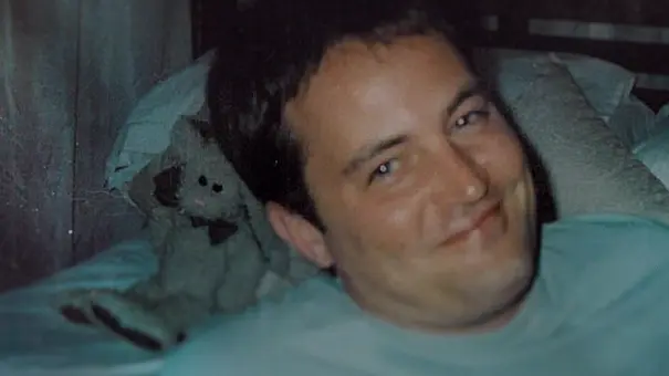 Matthew Perry liegt lächelnd in einem hellblauen Oberteil auf Kissen. Neben seinem Kopf sitzt ein graues Stofftier. - Foto: © 2026 Netflix, Inc. Matthew Perry liegt lächelnd in einem hellblauen Oberteil auf Kissen. Neben seinem Kopf sitzt ein graues Stofftier. - Foto: © 2026 Netflix, Inc.