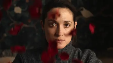 Schauspielerin Friederike Becht in ihrer Rolle als Lisa Konrad im grauen Rollkragenpullover blickt direkt in die Kamera. Unscharfe rote Blütenblätter schweben im Vordergrund. - Foto: Netflix Schauspielerin Friederike Becht in ihrer Rolle als Lisa Konrad im grauen Rollkragenpullover blickt direkt in die Kamera. Unscharfe rote Blütenblätter schweben im Vordergrund. - Foto: Netflix