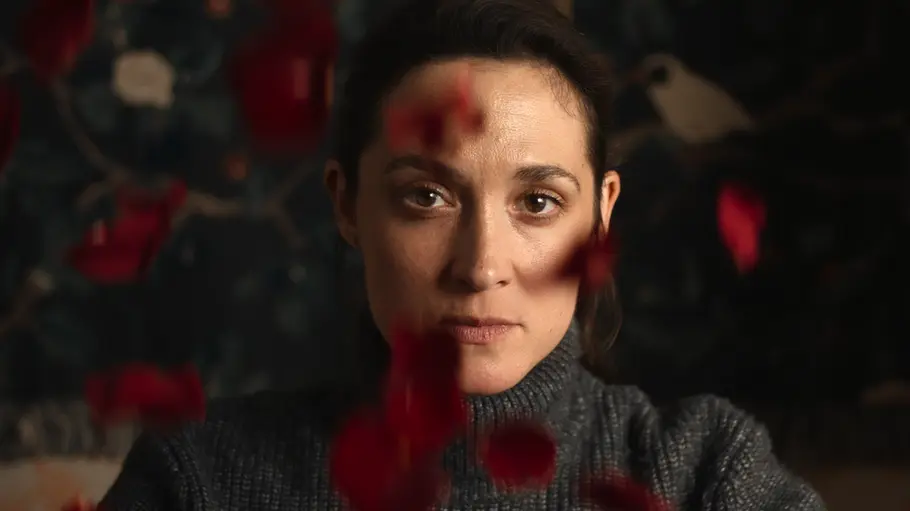 „Die Falle“ auf Netflix. Schauspielerin Friederike Becht in ihrer Rolle als Lisa Konrad im grauen Rollkragenpullover blickt direkt in die Kamera. Unscharfe rote Blütenblätter schweben im Vordergrund.
