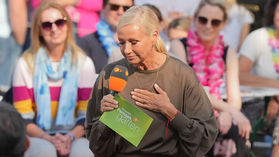 Die „Fernsehgarten“-Saison ist eigentlich vorbei, doch mit „Fernsehgarten on Tour“ geht es noch weiter. Andrea Kiewel hält ein ZDF-Mikrofon und eine grüne Moderationskarte vor Publikum. Ihre andere Hand liegt auf der Brust.