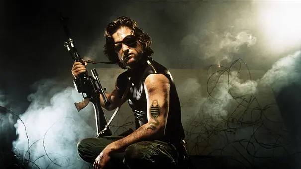Kurt Russell posiert als Snake Plissken mit einer Schusswaffe vor Stacheldraht im Nebel. - Foto:  IMAGO / Allstar