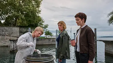 Mordufer - Kurzes Glück: Sarah Hertweg (Anna Gesa-Raija Lappe), Doro Beitinger (Franziska Weisz) und Chiara Locatelli (Maria Wördemann) stehen am Ufer des Bodensees vor einem offenen Fass. Hertweg trägt einen weißen Overall und weiße Handschuhe, mit der rechten Hand hält sie eine Spritze über das Fass. Beitinger und Locatelli beobachten sie dabei. - Foto: ZDF/Patrick Pfeiffer Konstanz