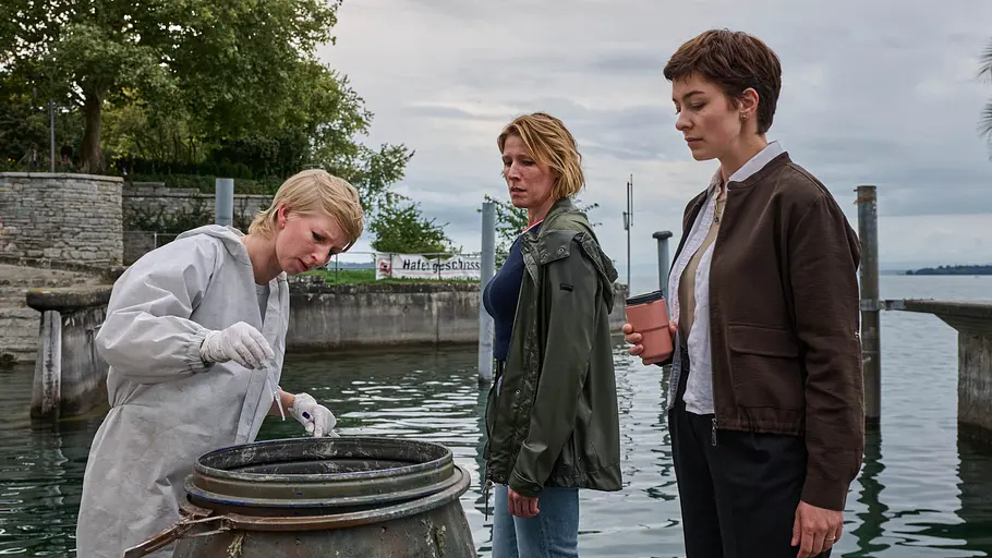 Die Kommissarinnen Doro Beitinger (Franziska Weisz, M.) und Chiara Locatelli (Maria Wördemann, r.) werden in ihrem zweiten gemeinsamen Fall mit einer Wasserleiche konfrontiert. Mordufer - Kurzes Glück: Sarah Hertweg (Anna Gesa-Raija Lappe), Doro Beitinger (Franziska Weisz) und Chiara Locatelli (Maria Wördemann) stehen am Ufer des Bodensees vor einem offenen Fass. Hertweg trägt einen weißen Overall und weiße Handschuhe, mit der rechten Hand hält sie eine Spritze über das Fass. Beitinger und Locatelli beobachten sie dabei.
