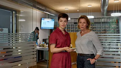 Mordufer - Die große Gier: Chiara Locatelli (Maria Wördemann) und Doro Beitinger (Franziska Weisz) stehen in einem Büro und blicken zur Seite. Locatelli hält ein Smartphone in ihrer rechten Hand. Im Hintergrund steht Katrin Glaunsinger (Beate Constanze Pitronik) an einem Schreibtisch und telefoniert mit einem Festnetztelefon. - Foto:  ZDF / Patrick Pfeiffer