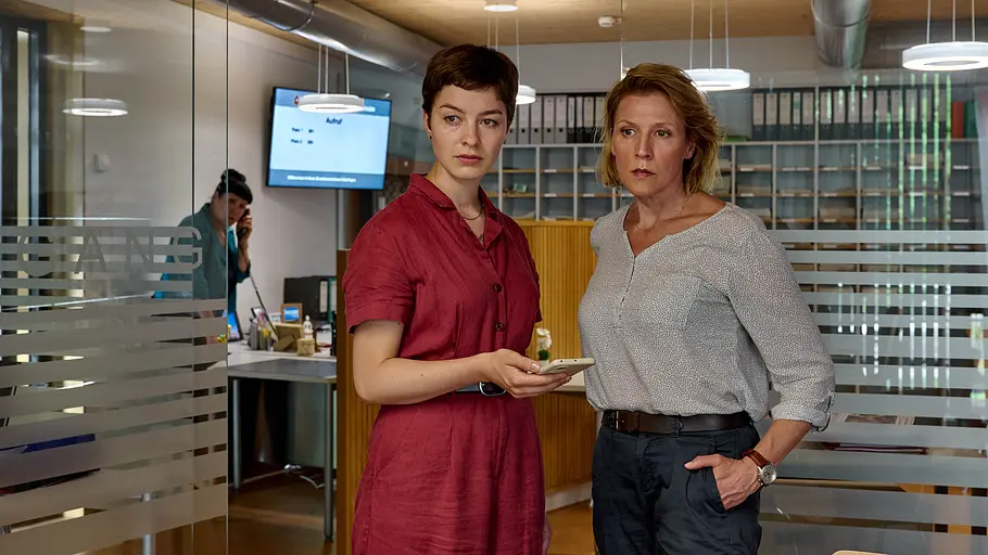 Die Kriminalkommissarinnen Chiara Locatelli (Maria Wördemann, M.) und Doro Beitinger (Franziska Weisz, r.) ermitteln im Einwohnermeldeamt. Katrin Glaunsinger (Beate Constanze Pitronik, l.) kann offenbar wenig zur Aufklärung des Mordes an ihrer Mitarbeiterin beitragen. Mordufer - Die große Gier: Chiara Locatelli (Maria Wördemann) und Doro Beitinger (Franziska Weisz) stehen in einem Büro und blicken zur Seite. Locatelli hält ein Smartphone in ihrer rechten Hand. Im Hintergrund steht Katrin Glaunsinger (Beate Constanze Pitronik) an einem Schreibtisch und telefoniert mit einem Festnetztelefon.