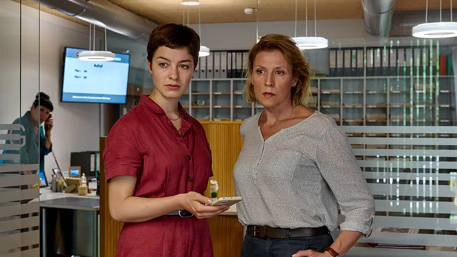 Die Kriminalkommissarinnen Chiara Locatelli (Maria Wördemann, M.) und Doro Beitinger (Franziska Weisz, r.) Mordufer - Die große Gier: Chiara Locatelli (Maria Wördemann) und Doro Beitinger (Franziska Weisz) stehen in einem Büro und blicken zur Seite. Locatelli hält ein Smartphone in ihrer rechten Hand. Im Hintergrund steht Katrin Glaunsinger (Beate Constanze Pitronik) an einem Schreibtisch und telefoniert mit einem Festnetztelefon.