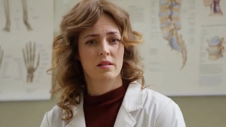 DIE LANDARZTPRAXIS-Vorschau FOLGE 88 Mittwoch 28. Oktober Ella trägt einen weißen Kittel über einem dunkelroten Oberteil. Hinter ihr hängen anatomische Schaubilder an der Wand.