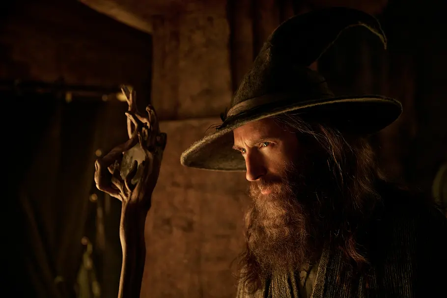 Gandalf stellt sich der Dunkelheit Gandalf trägt einen breiten, spitzen Hut und einen langen Bart. Neben ihm steht ein knorriger Holzstab im gedämpften Licht.