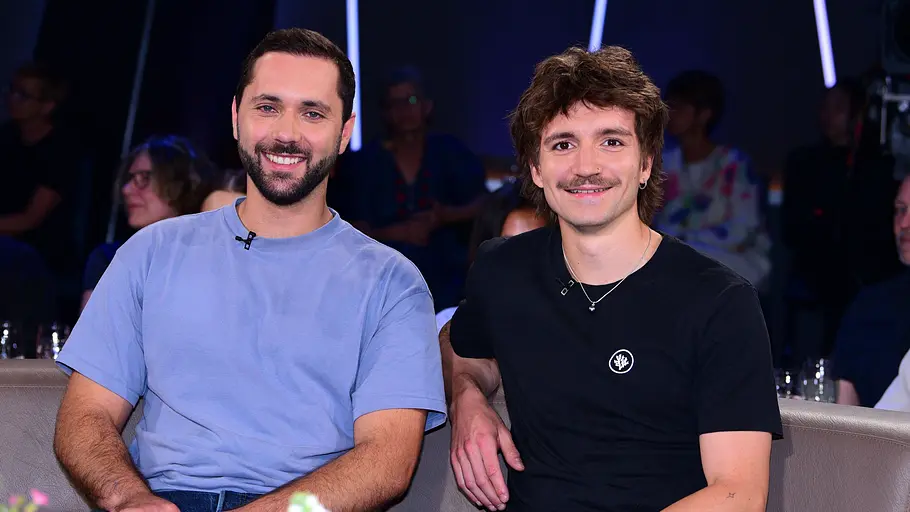 Die „Segeljungs“ Die Content Creator „Segeljungs“ sitzen gemeinsam auf einem Sofa in einem TV-Studio und lächeln in die Kamera.