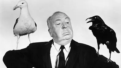 Alfred Hitchcock posiert mit zwei Vögeln. Links ist eine Möwe, rechts ist ein Rabe. - Foto:  IMAGO / Everett Collection