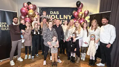 Mehrere Erwachsene und Kinder stehen gemeinsam vor einer schwarzen Fotowand mit der Aufschrift „Das Wollny Quiz“. Goldene, schwarze und rosafarbene Luftballons rahmen die Szene ein. Einige Kinder haben geschminkte Gesichter, während die Gruppe lächelnd in die Kamera blickt. - Foto: RTLZWEI