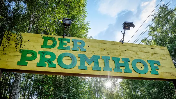 Ein gelbes Holzschild mit der grünen Aufschrift „DER PROMIHOF“ hängt zwischen Bäumen. Darüber sind zwei Strahler angebracht. - Foto: RTLZWEI Ein gelbes Holzschild mit der grünen Aufschrift „DER PROMIHOF“ hängt zwischen Bäumen. Darüber sind zwei Strahler angebracht. - Foto: RTLZWEI
