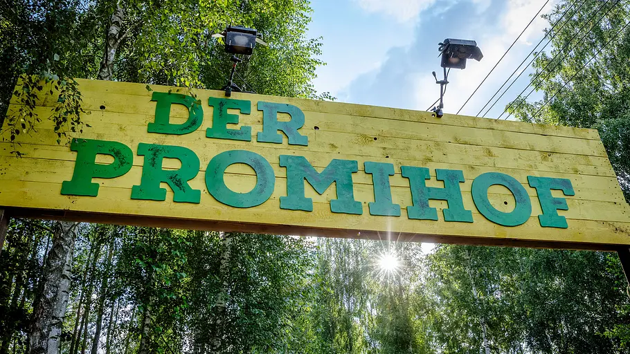 Die zweite Staffel von „Der Promihof“ steht bevor Ein gelbes Holzschild mit der grünen Aufschrift „DER PROMIHOF“ hängt zwischen Bäumen. Darüber sind zwei Strahler angebracht.