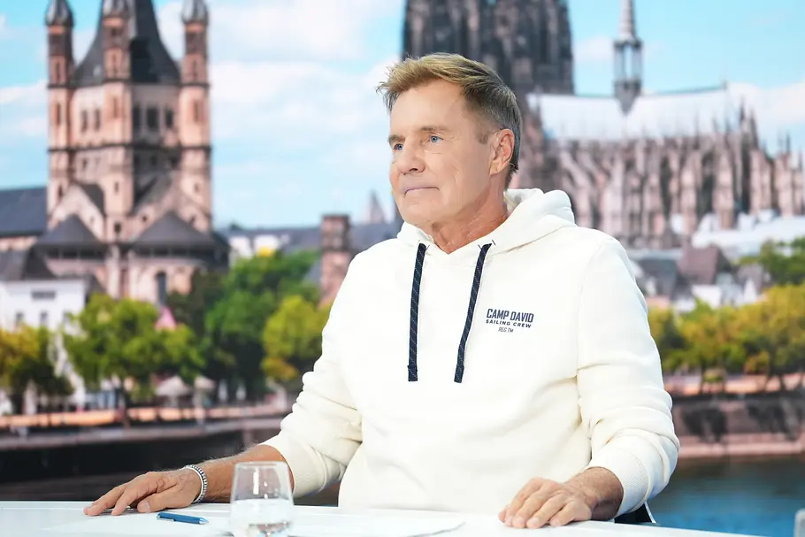 Dieter Bohlen sitzt im DSDS-Studio vor einer Köln-Kulisse mit Dom im Hintergrund, trägt weißen Hoodie und sitzt an einem Tisch mit Wasserglas.