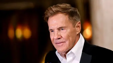 Mann in schwarzem Smoking mit weißem Hemd gibt ein Interview und spricht in ein Mikrofon, im Hintergrund warmes Bühnenlicht. - Foto: IMAGO / Panama Pictures