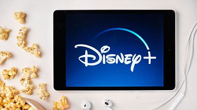 Ein Tablet, auf dem das Disney+-Logo zu sehen ist. Daneben liegt Popcorn. - Foto: IMAGO / Dreamstime