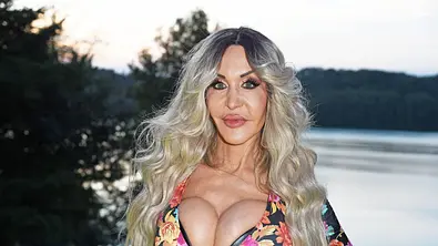 Dolly Buster posiert mit langen blonden Haaren und gemustertem Oberteil vor einem See. - Foto:  IMAGO / Gartner